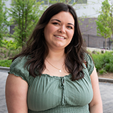 Morgan Karcher stands outside, smiling, in green dress, with trees and concrete patio in background